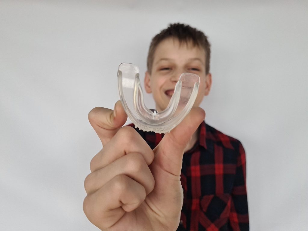 A,Boy,Smiles,While,Holding,A,Clear,Mouthguard,In,Front