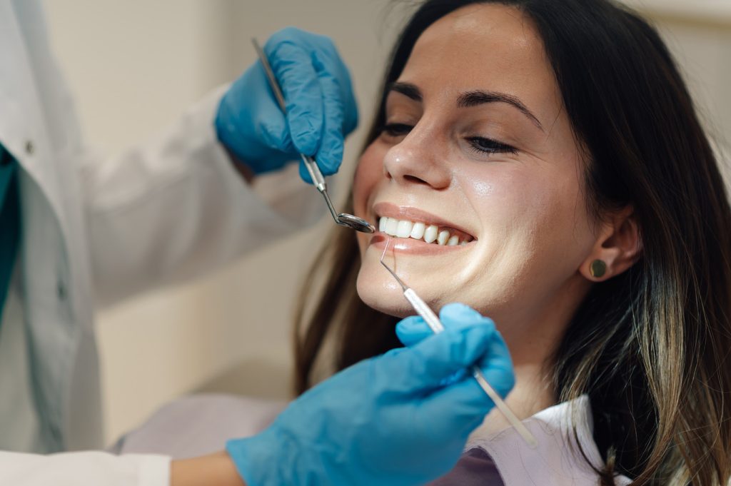 Dentist,Wearing,Blue,Gloves,Examining,Teeth,Of,Smiling,Young,Woman