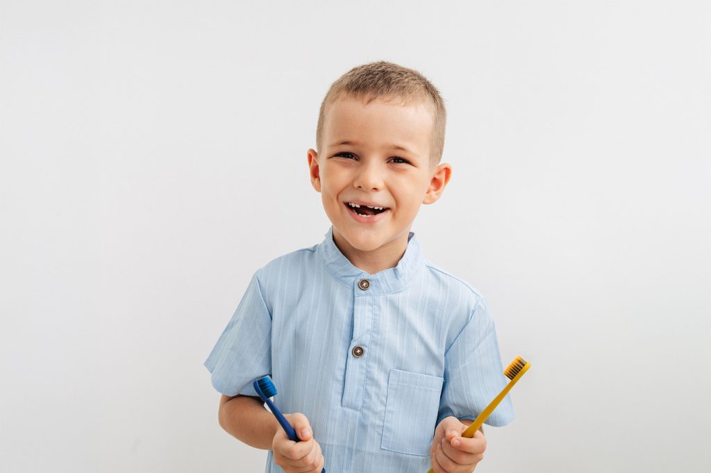 Child,Holding,Yellow,And,Blue,Toothbrushes,,Smiling,With,A,Missing
