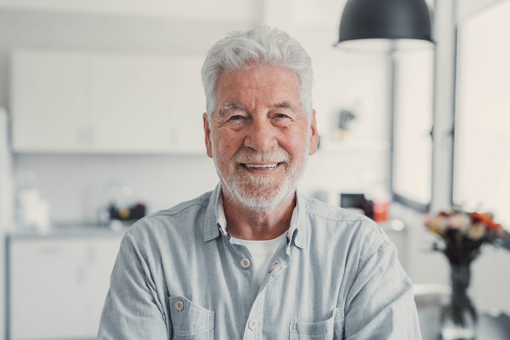 Elderly,Man,Standing,In,Living,Room,Smiling,At,The,Camera.